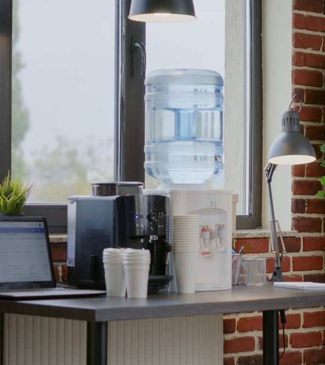Close up of water dispenser and coffee machine on table for people working in office. Water cooler on desk with cups and equipment used for liquid refreshment and beverage on break.